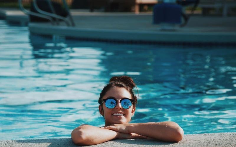 woman with sunglasses, in a pool, rests her arms on the ledge and smiles